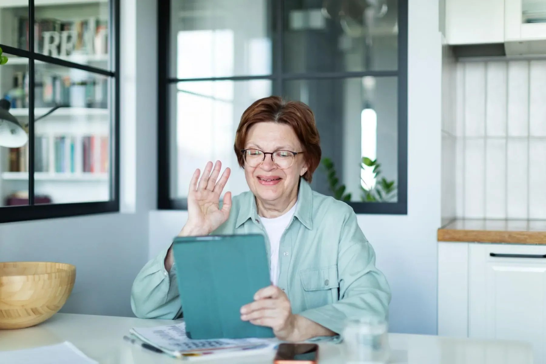 An older woman smiling and waving at her tablet during a video call at home