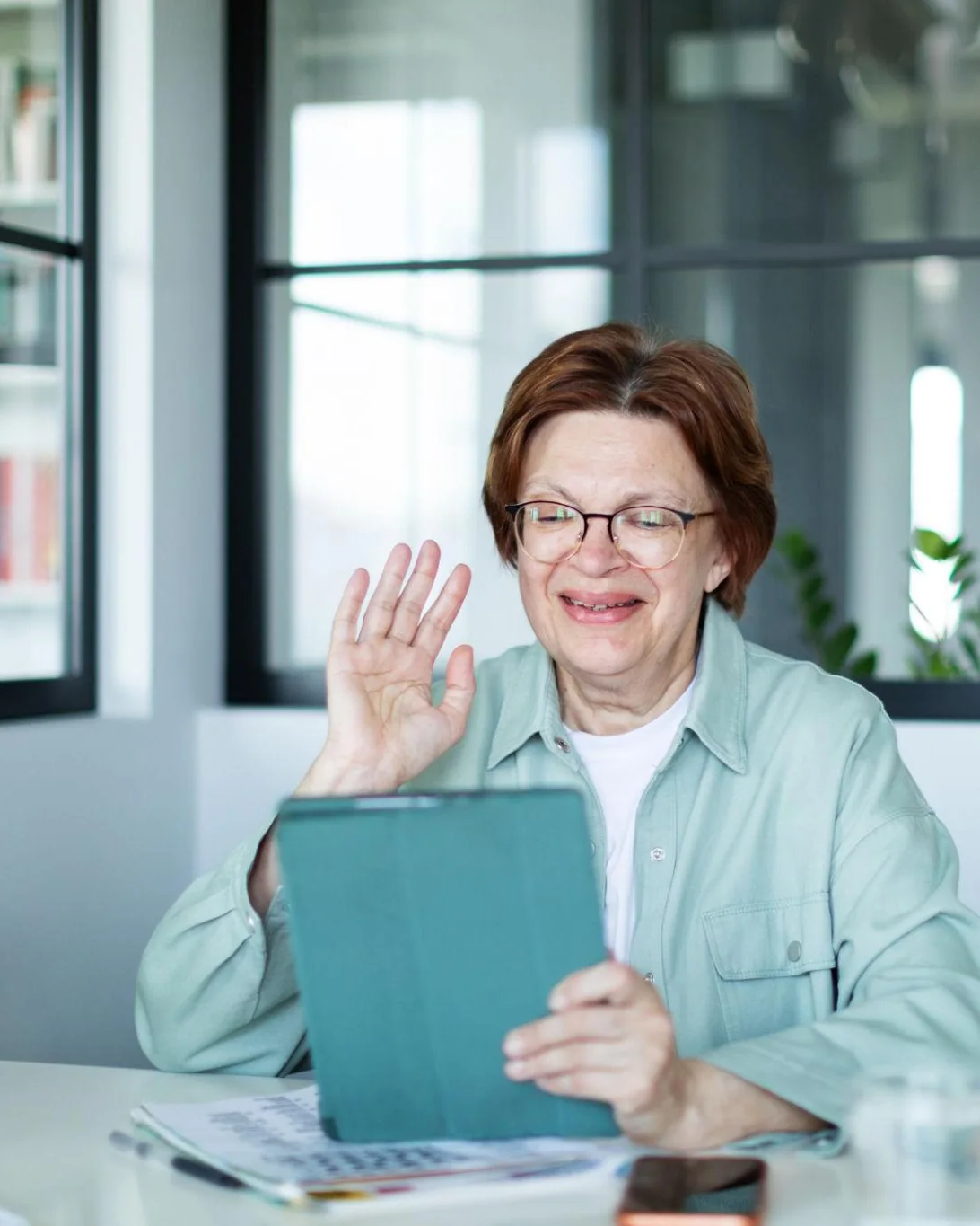 An older woman smiling and waving at her tablet during a video call at home