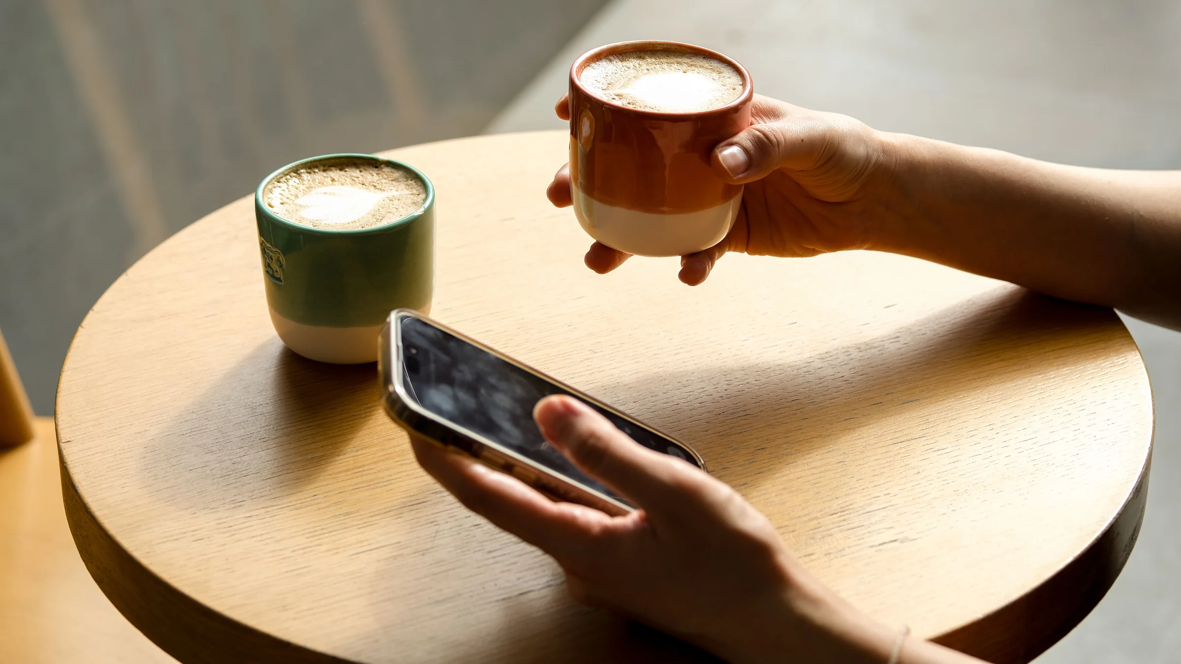 Two cups of coffee on a small round table, with someone holding a cup and a phone