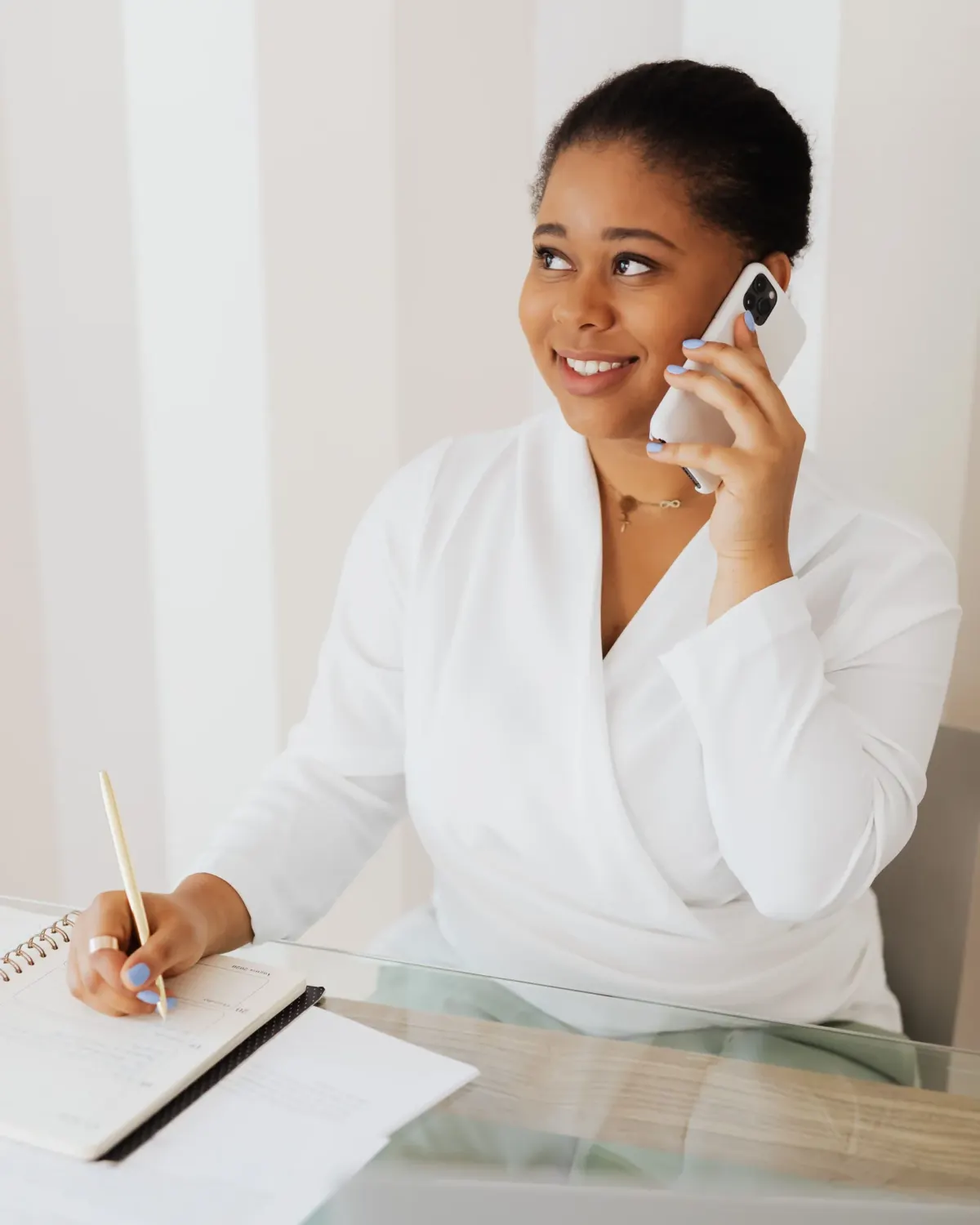 A woman smiling on the phone at her desk, writing in a notebook