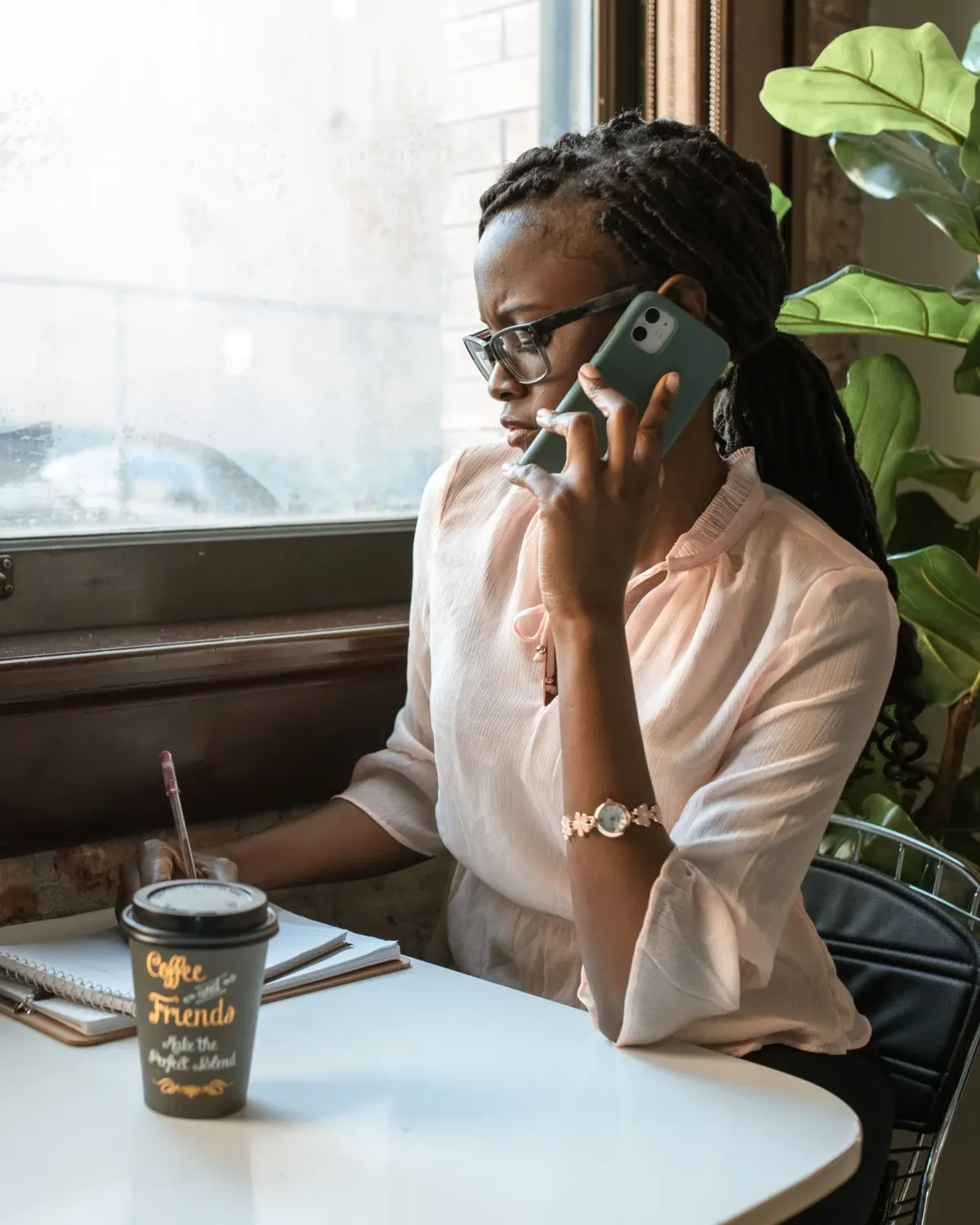 A woman by a window talking on her phone while writing in a notebook